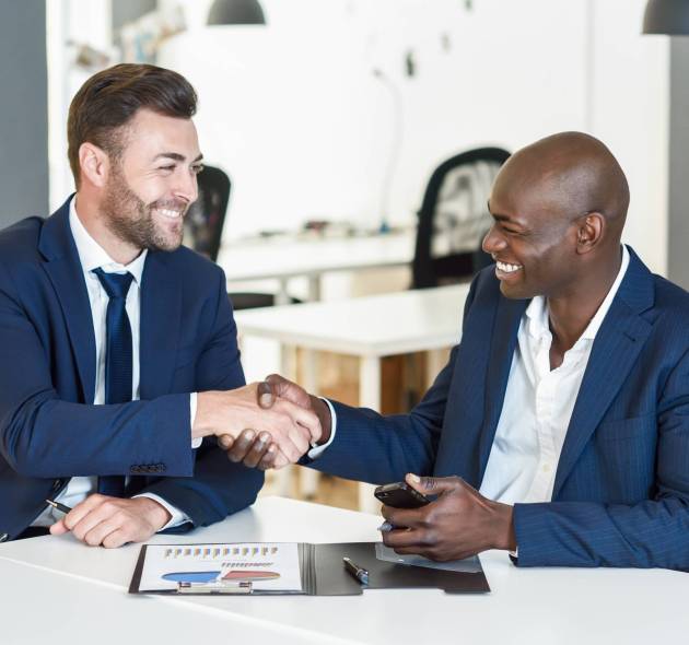 Black businessman shaking hands with a caucasian one wearing suit in a office. Two smiling men wearing blue suits working in an office with white furniture