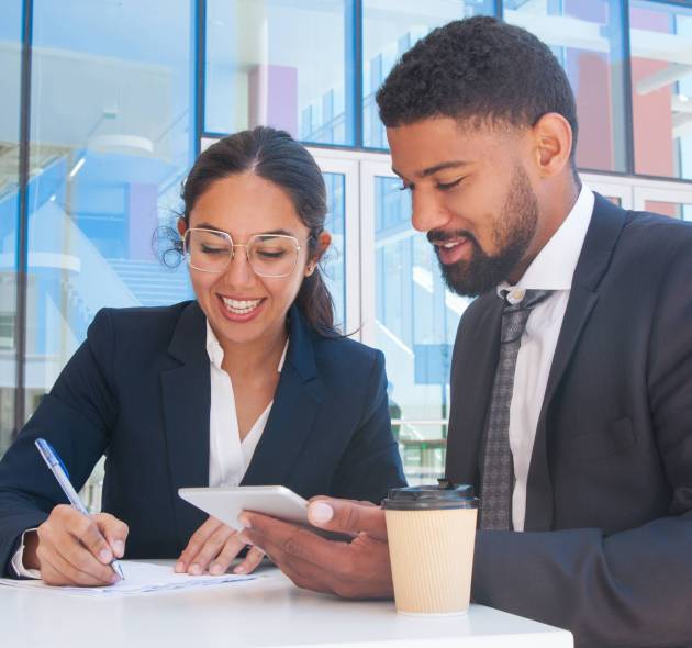 Smiling business people working and using tablet in outdoor cafe. Business man and woman wearing formal clothes and sitting with building glass wall in background. Partners concept.